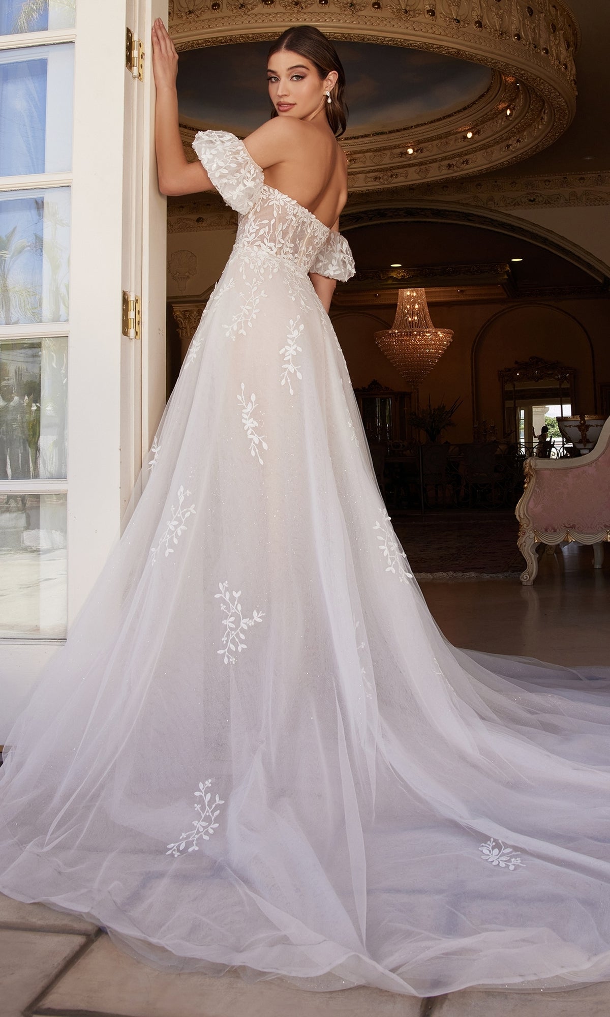 A woman wearing the Puff-Sleeve Long White Wedding Dress A1014, featuring floral lace details and removable puff sleeves, stands by an open door in an ornate room, glancing back over her shoulder.
